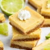 Three key lime bars are stacked, topped with whipped cream, with more bars and lime wedges scattered around on a white surface.