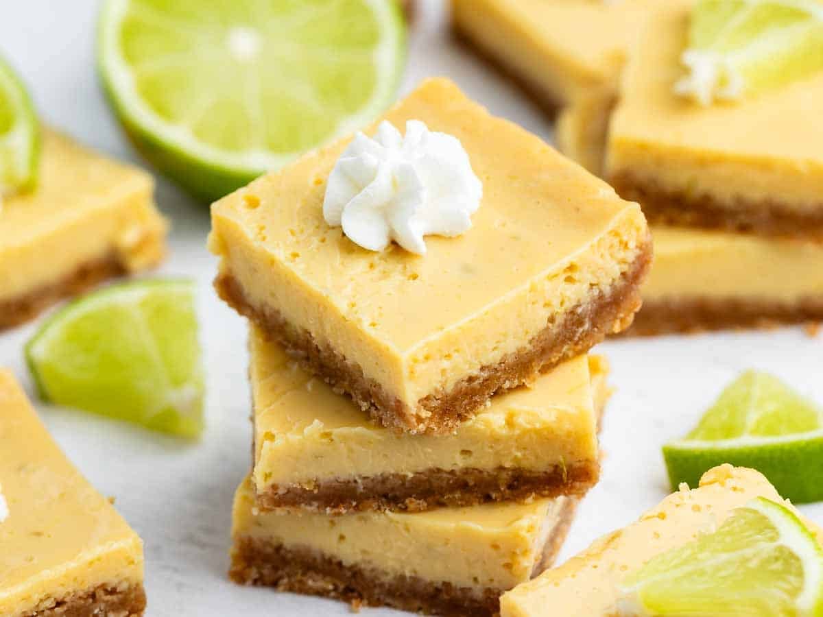 Three key lime bars are stacked, topped with whipped cream, with more bars and lime wedges scattered around on a white surface.