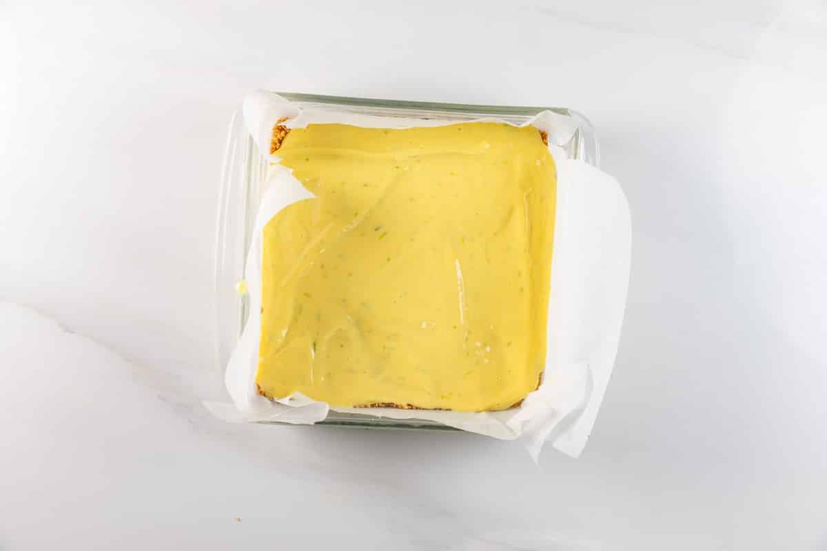 A glass baking dish lined with parchment paper holds a rectangular dessert topped with a smooth layer of yellow filling, possibly lemon or custard, on a light marble surface.
