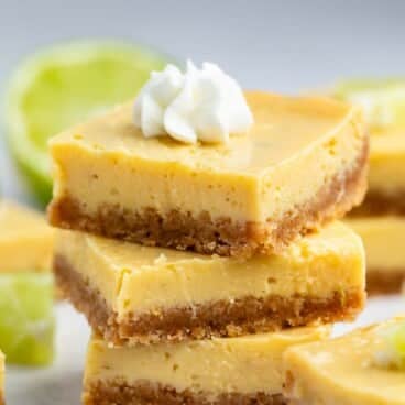 Three stacked key lime bars with a graham cracker crust, topped with a dollop of whipped cream, with a lime half and more bars blurred in the background.