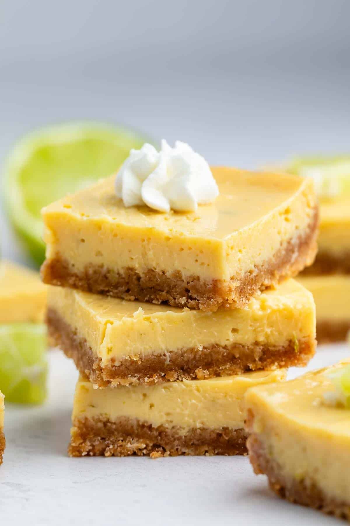 Three stacked key lime bars with a graham cracker crust, topped with a dollop of whipped cream, with a lime half and more bars blurred in the background.