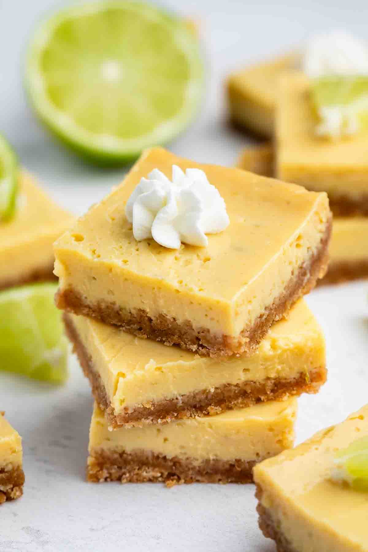 Three stacked key lime bars with a graham cracker crust, topped with whipped cream, are in focus. More bars and a sliced lime are in the background on a light surface.