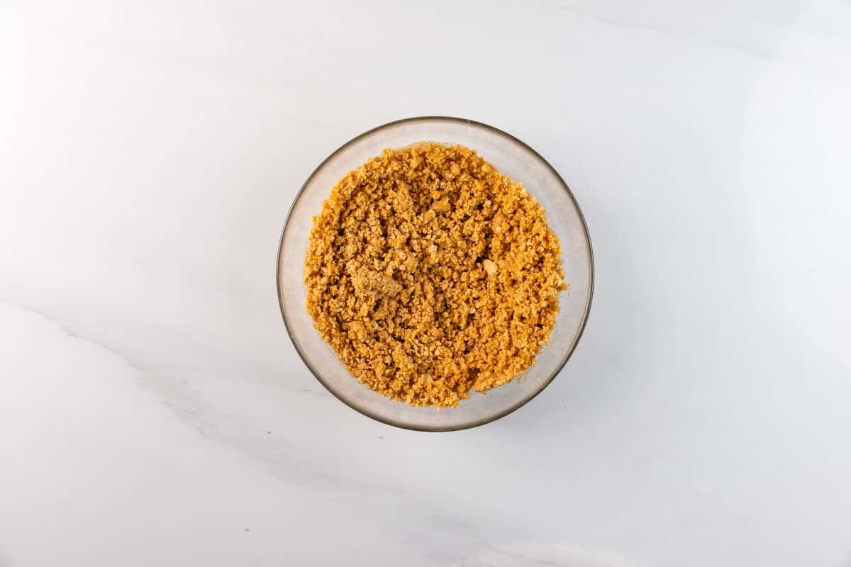 A small glass bowl filled with finely crumbled graham cracker crust sits on a white marble surface.