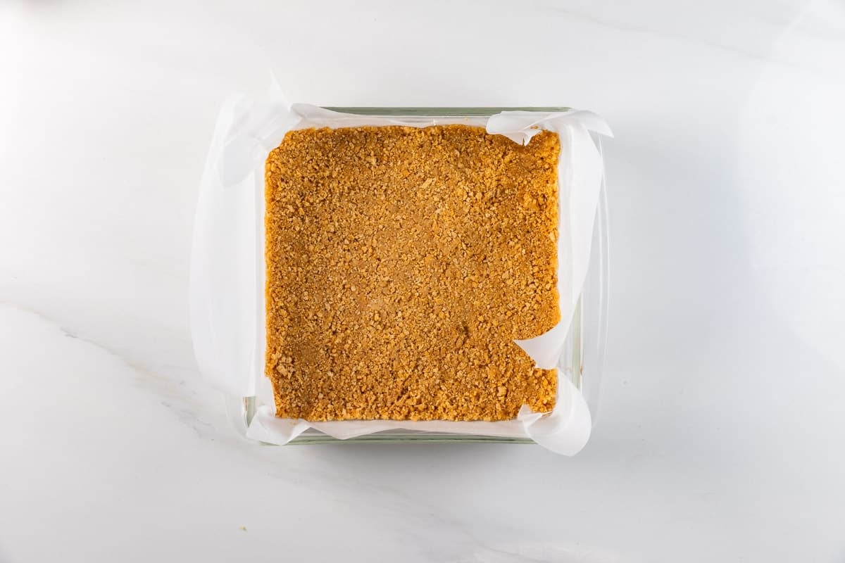 A square glass baking dish lined with parchment paper holds a flat, golden-brown graham cracker crust on a white marble surface.