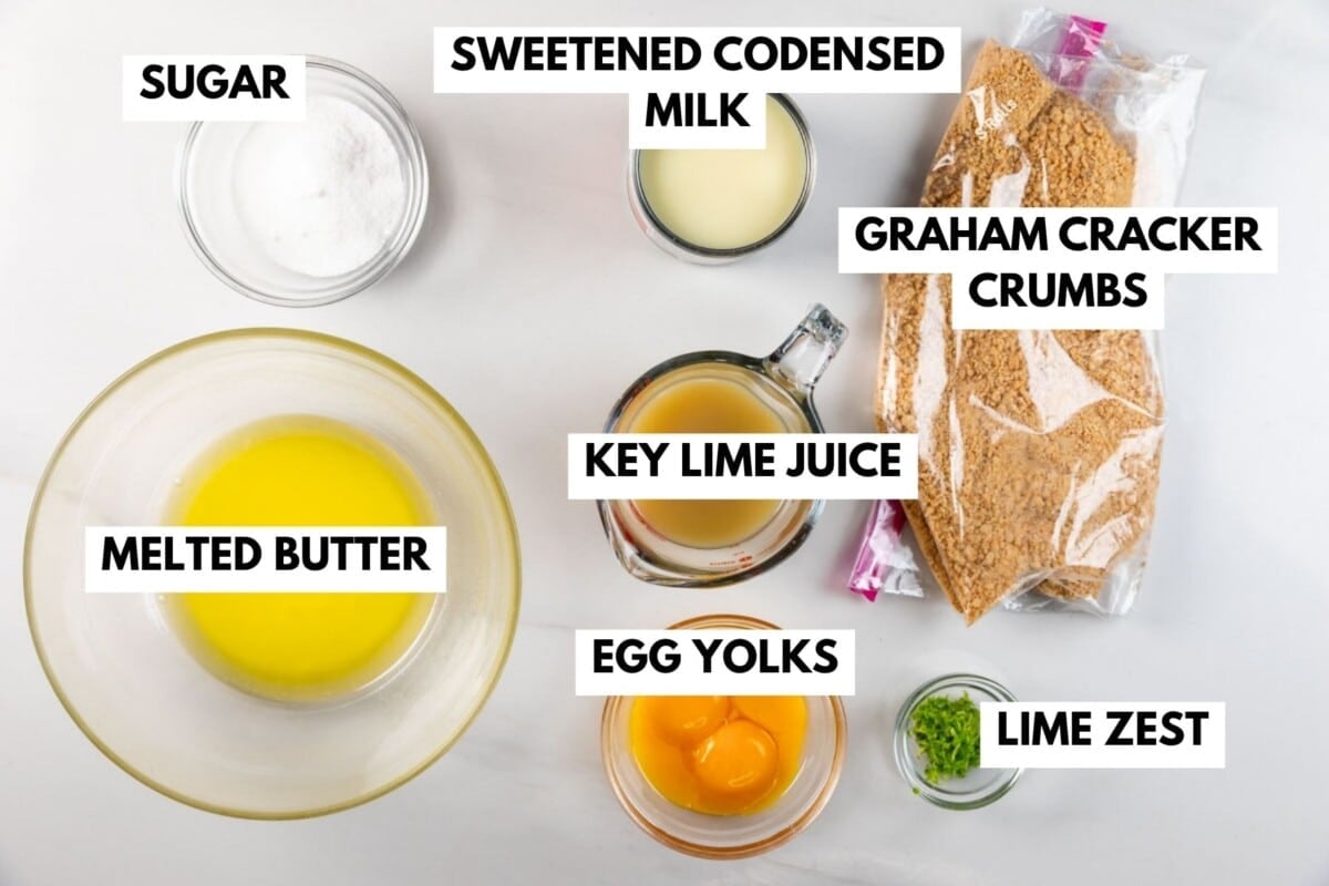 Top view of labeled ingredients for key lime pie: sugar, sweetened condensed milk, graham cracker crumbs, melted butter, key lime juice, egg yolks, and lime zest, all arranged on a white surface.