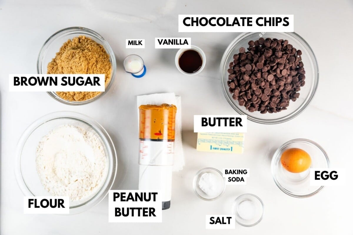 Bowls on a counter labeled with ingredients: brown sugar, flour, peanut butter, chocolate chips, butter, milk, vanilla, an egg, baking soda, and salt, for making cookies.