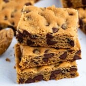 Three stacked chocolate chip cookie bars on a white surface, with more bars and a spoonful of peanut butter nearby.