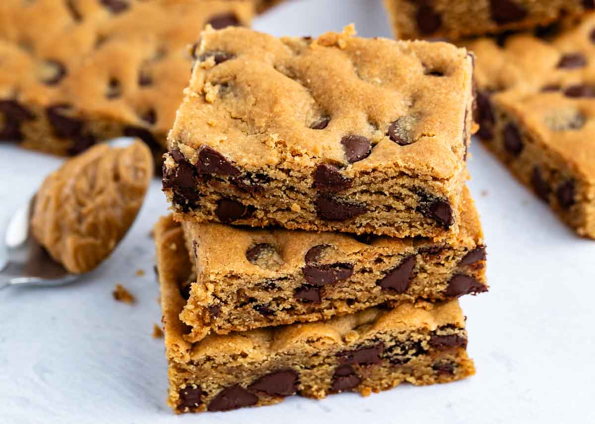 Three stacked chocolate chip cookie bars on a white surface, with more bars and a spoonful of peanut butter nearby.