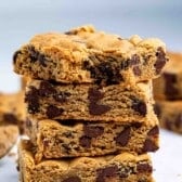 A stack of four thick chocolate chip cookie bars sits on a white surface, with more cookie bars and crumbs scattered around them.