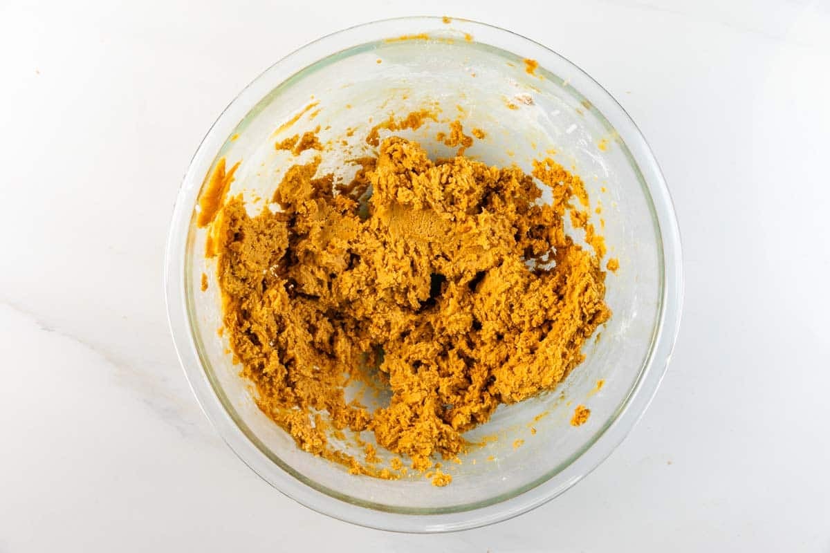 A glass bowl filled with orange-brown cookie dough sits on a white surface.