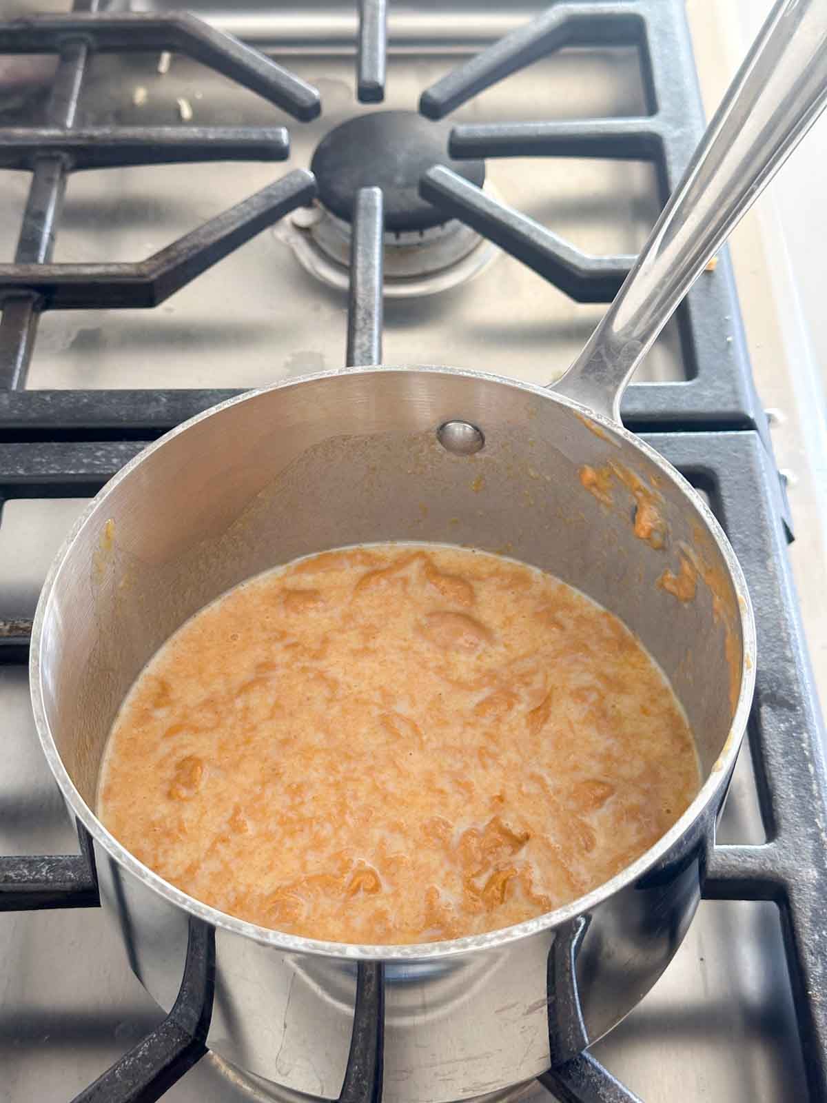 A stainless steel pot with bubbling, light brown oatmeal mixture sits on a gas stove with black grates. A metal spoon handle rests on the side of the pot.