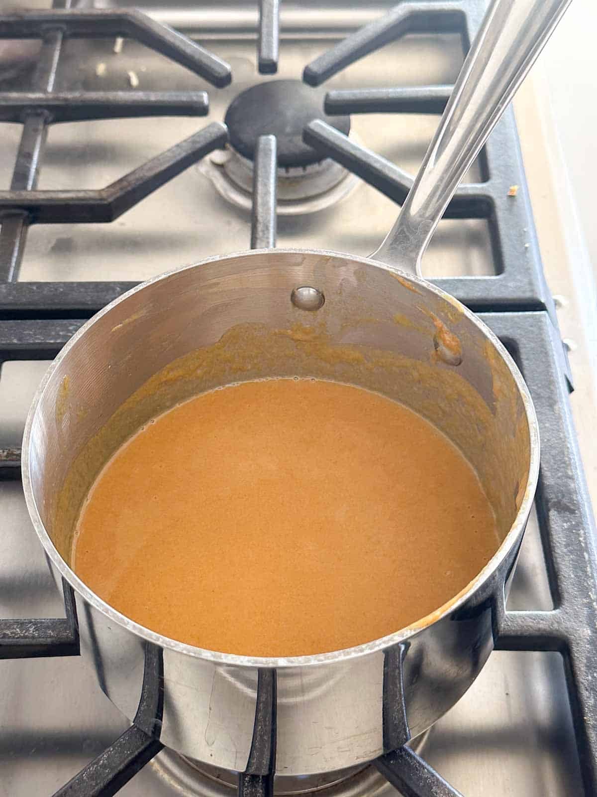 A stainless steel saucepan with a light brown sauce simmering on a gas stove. The sauce covers the bottom and sides of the pan, and the stovetop grates are visible around it.