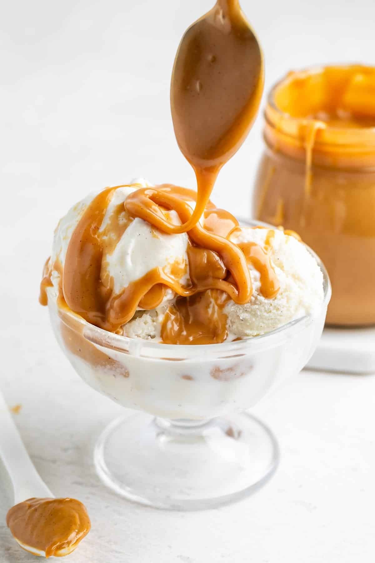 A spoon drizzles caramel sauce over vanilla ice cream in a glass bowl, with a jar of caramel sauce and a spoon resting nearby on a white surface.