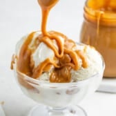 A glass bowl of vanilla ice cream is being topped with thick caramel sauce, with a jar of caramel sauce in the background.