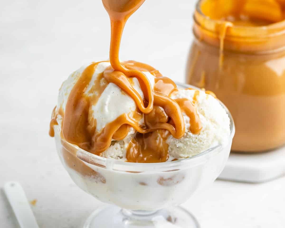 A glass bowl of vanilla ice cream is being topped with thick caramel sauce, with a jar of caramel sauce in the background.