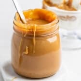 A jar of thick caramel sauce with a spoon inside, caramel dripping down the side, placed on a white surface with a dessert in the background.