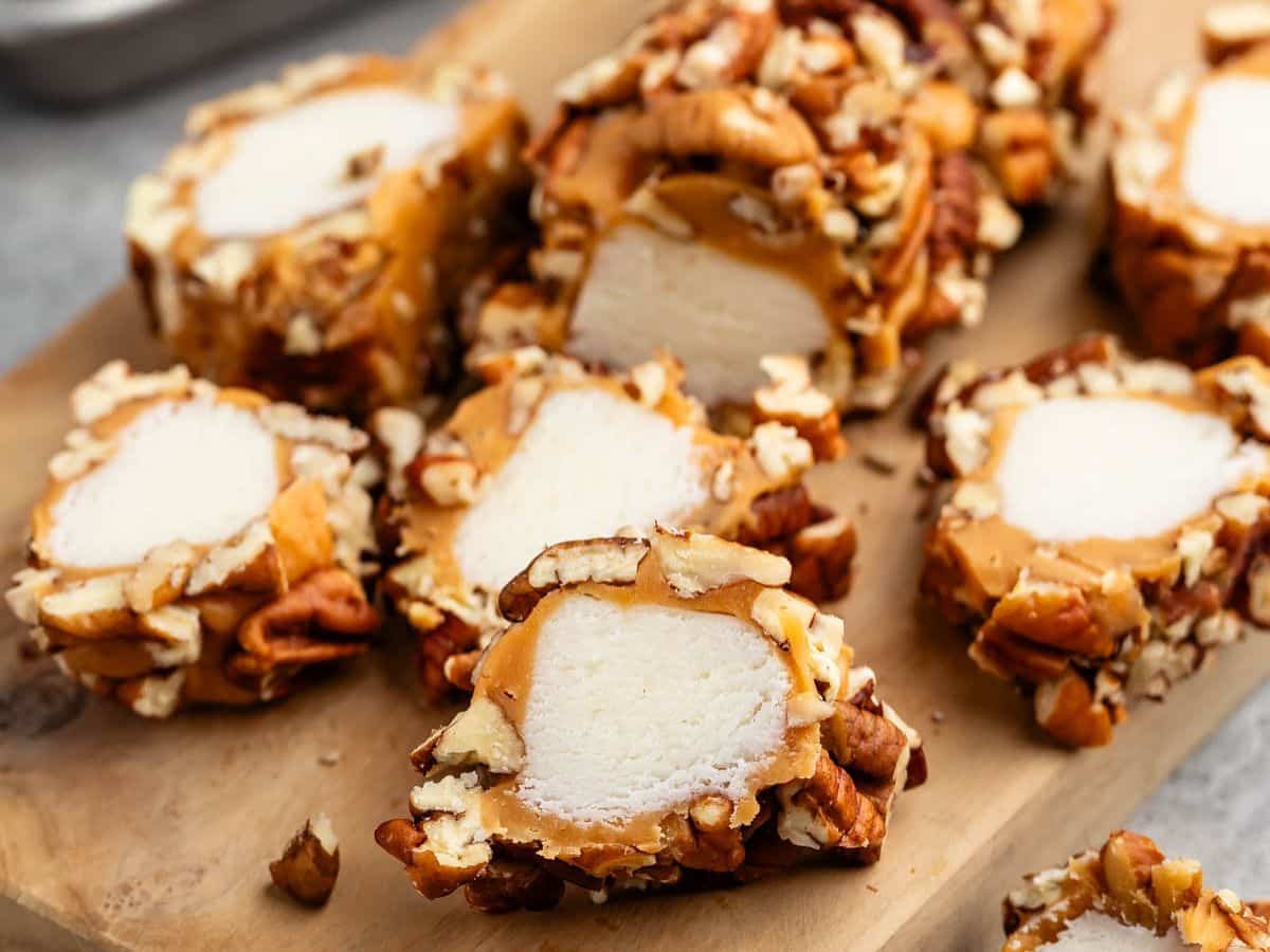 Sliced pieces of caramel-wrapped marshmallows coated in chopped pecans are arranged on a wooden board, showcasing their creamy, nutty texture and sweet, chewy appearance.