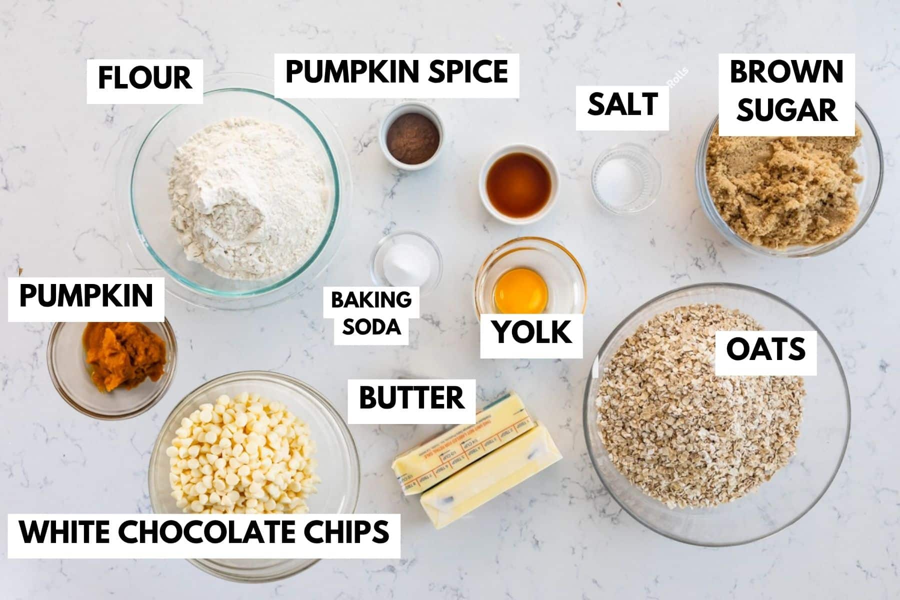 Bowls on a counter filled with labeled ingredients: flour, pumpkin spice, brown sugar, salt, pumpkin, baking soda, egg yolk, butter, oats, and white chocolate chips, arranged for baking.