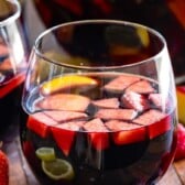 A close-up of glasses filled with red sangria, containing sliced strawberries, apples, lemons, and oranges. Fresh strawberries are placed beside the glasses on a wooden surface.