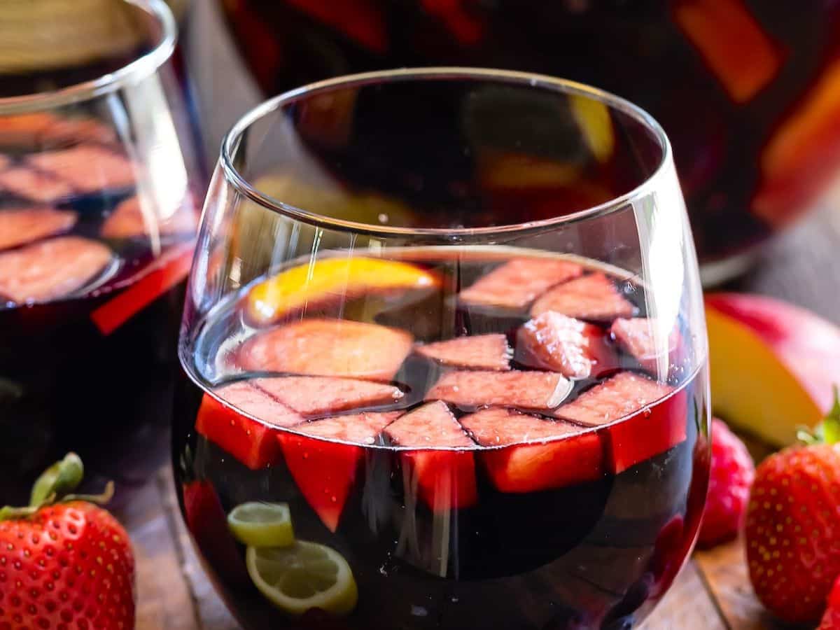 A close-up of glasses filled with red sangria, containing sliced strawberries, apples, lemons, and oranges. Fresh strawberries are placed beside the glasses on a wooden surface.