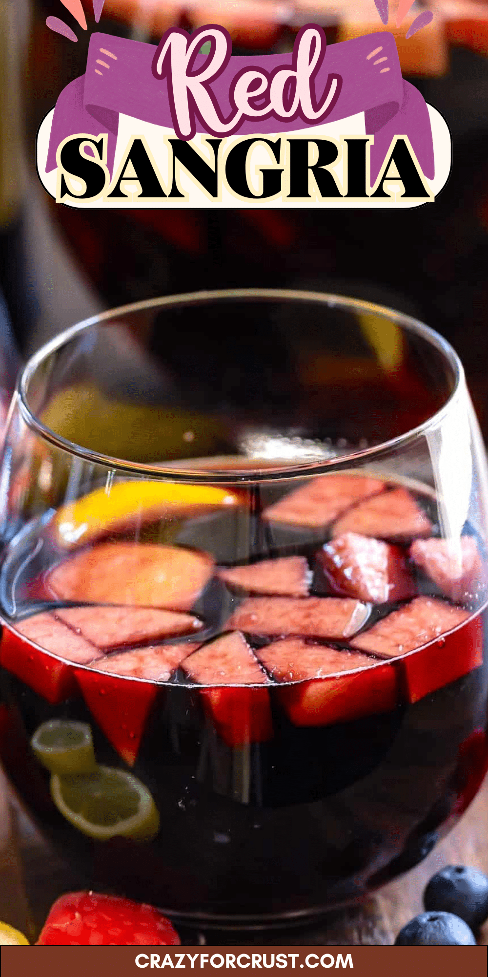 A close-up of a glass of red sangria filled with ice, fruit slices, and berries. The text Red Sangria appears at the top, and crazyforcrust.com is at the bottom.