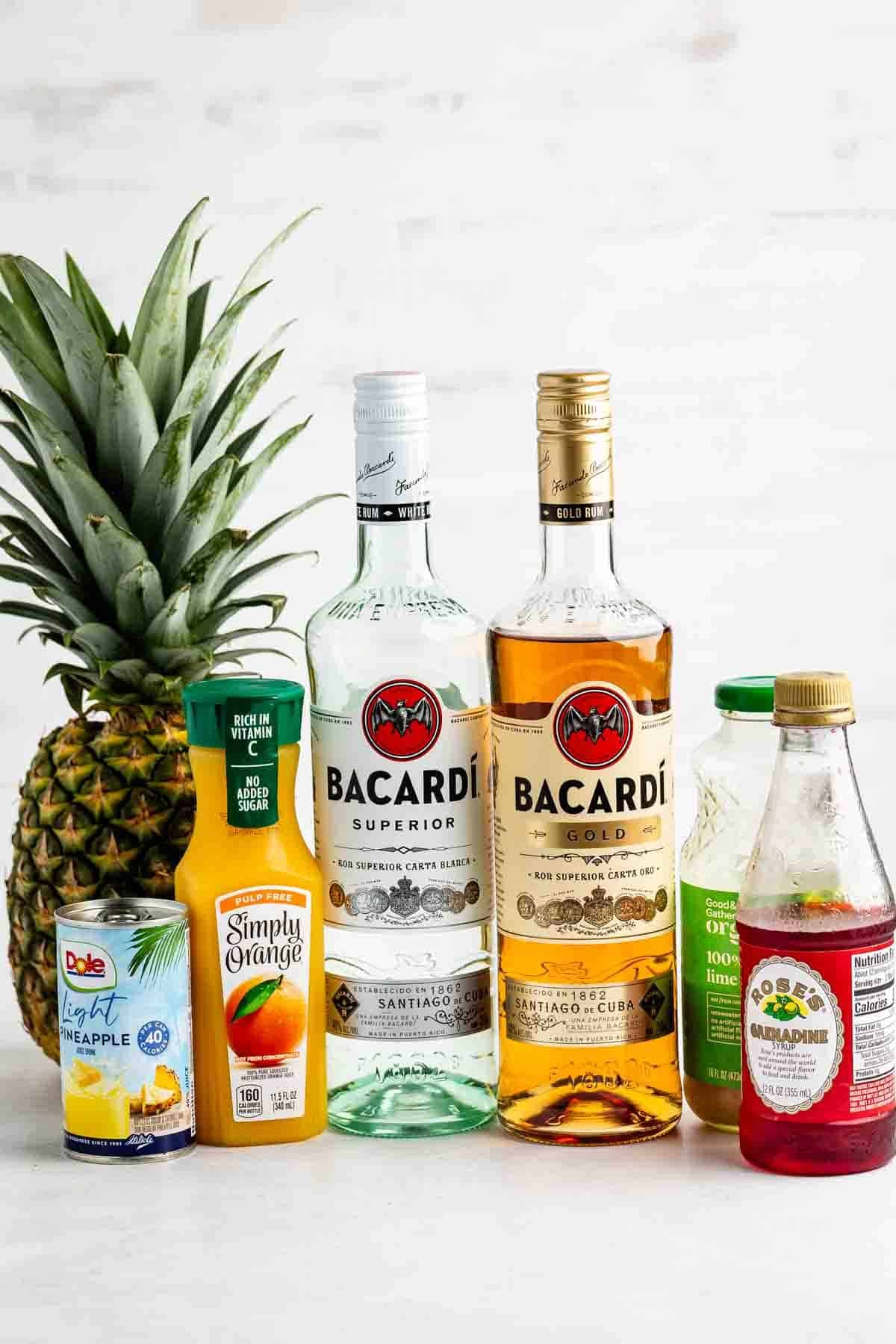Two bottles of Bacardi rum, a fresh pineapple, canned pineapple juice, orange juice, grenadine, and lime juice are arranged on a white surface with a light background.