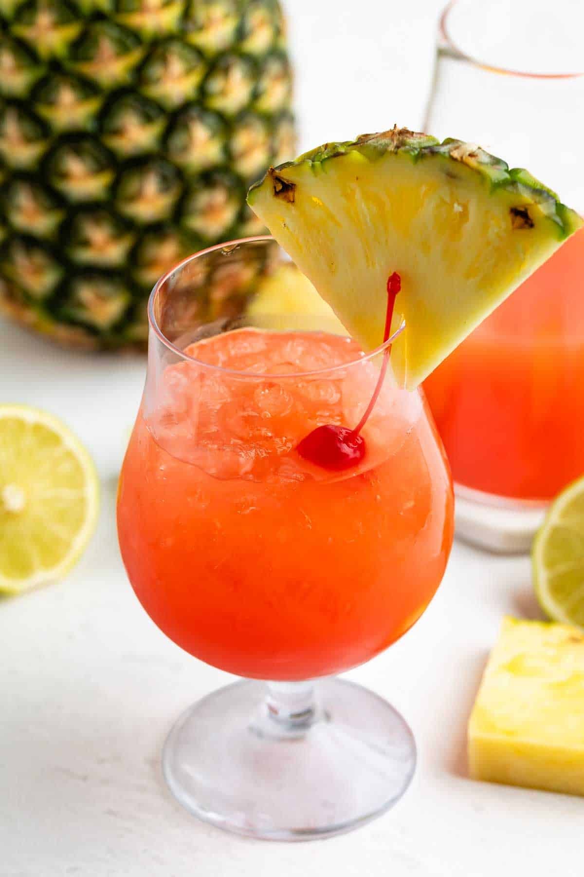 A tropical cocktail with orange-red liquid, crushed ice, a maraschino cherry, and a pineapple wedge garnish. Pineapple and lime slices are in the background.