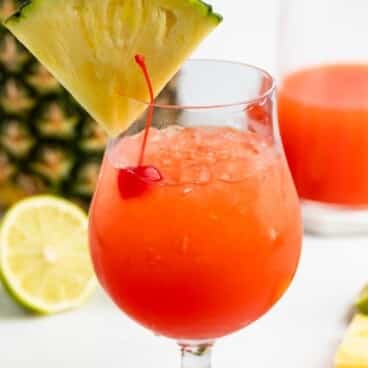 A bright red tropical drink in a curved glass, garnished with a pineapple slice and a cherry, with a lime slice, pineapple, and a glass of orange liquid in the background.