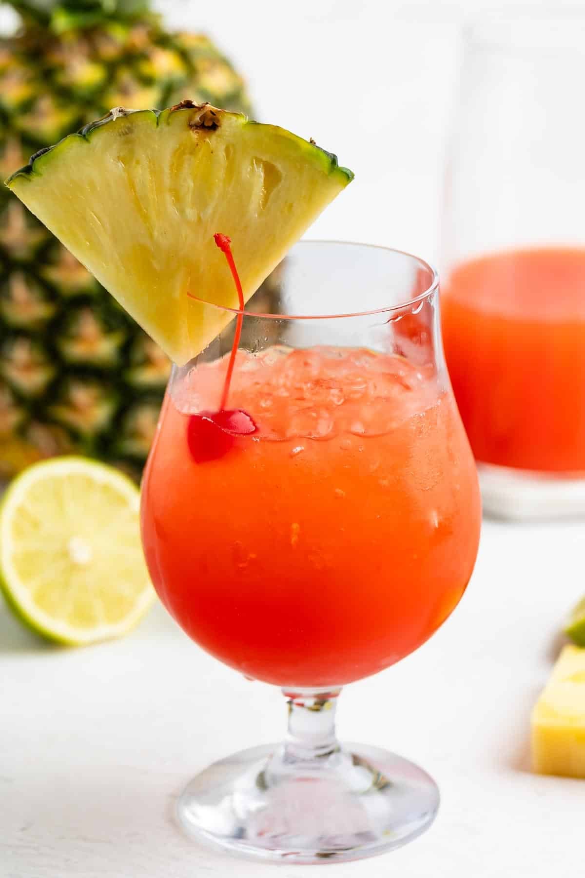 A bright red tropical drink in a curved glass, garnished with a pineapple slice and a cherry, with a lime slice, pineapple, and a glass of orange liquid in the background.