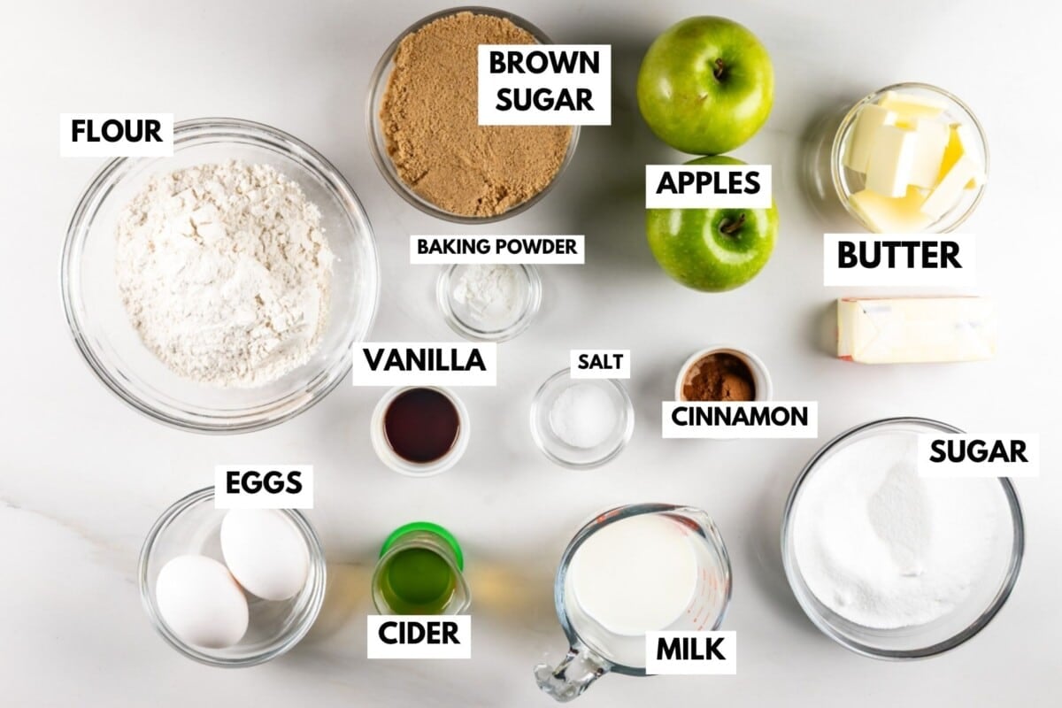 Ingredients for baking are arranged on a white surface, including bowls of flour, brown sugar, sugar, baking powder, salt, cinnamon, eggs, cider, vanilla, milk, butter, and two green apples. Labels identify each item.