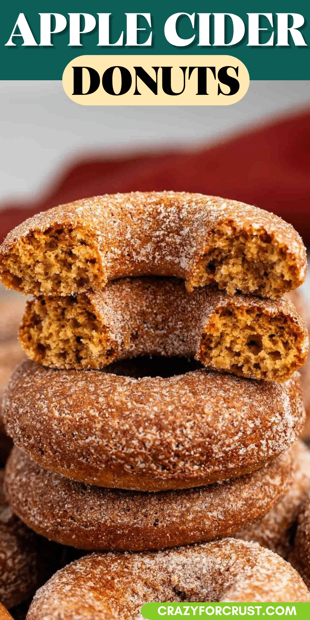 A stack of apple cider donuts, dusted with sugar, with one apple cider donut on top broken in half to show the soft inside. Text above reads Apple Cider Donuts.