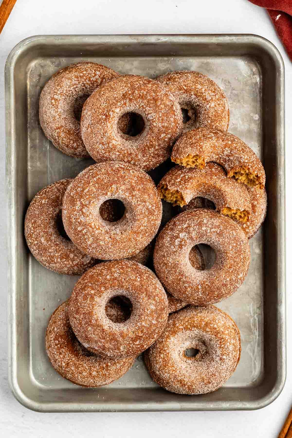 A metal tray filled with baked cinnamon sugar donuts, some stacked and two partially eaten, on a white surface.