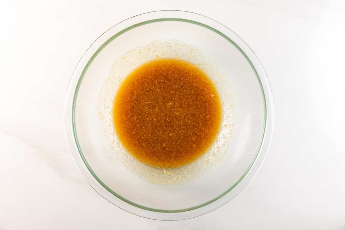 A glass bowl containing a light brown liquid mixture is placed on a white countertop. The mixture appears to be partially blended, with some grainy texture visible.