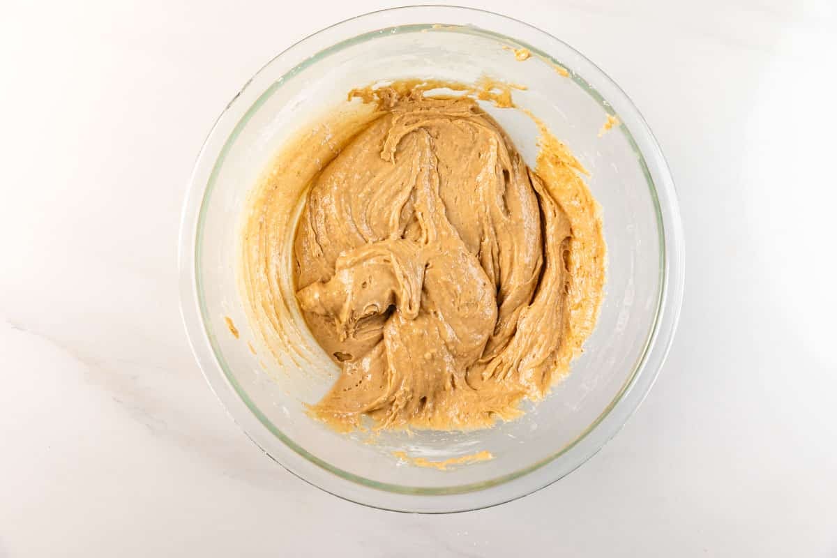 A glass bowl filled with creamy, light brown cookie dough sits on a white marble surface.