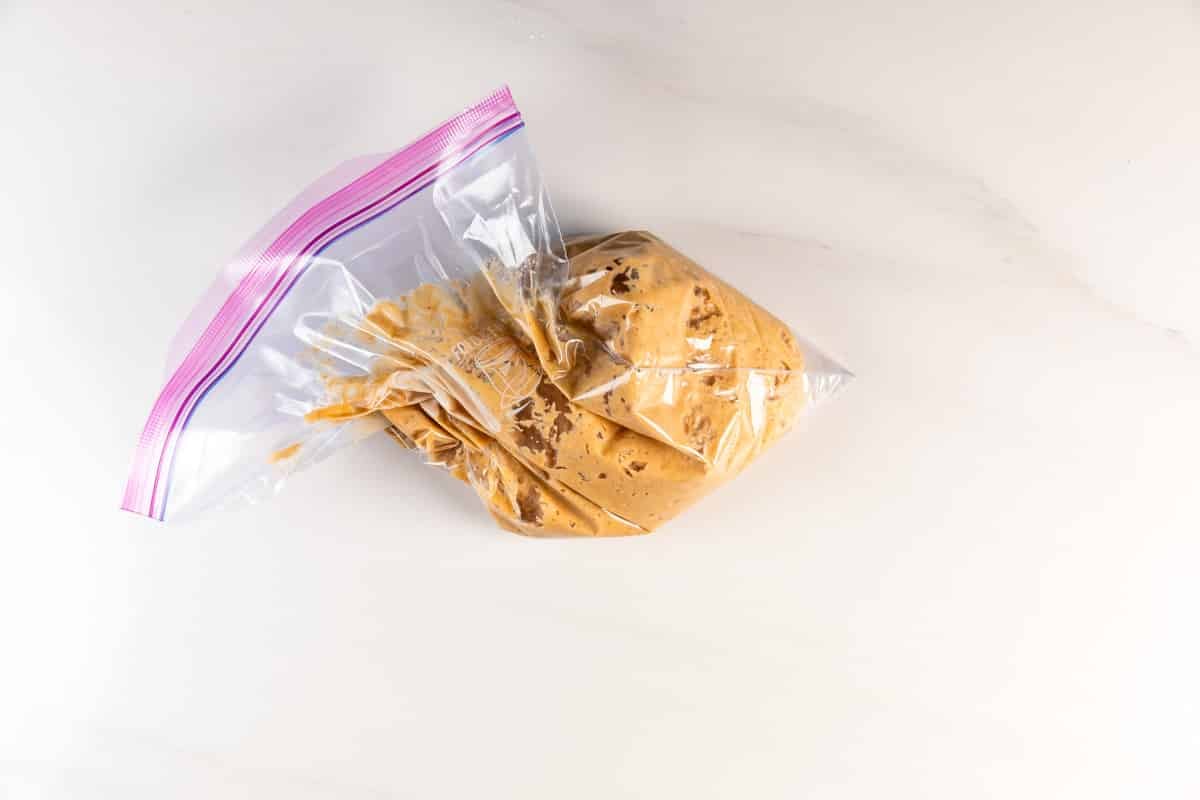 A clear plastic zip-top bag filled with light brown cookie dough rests on a white marble surface.