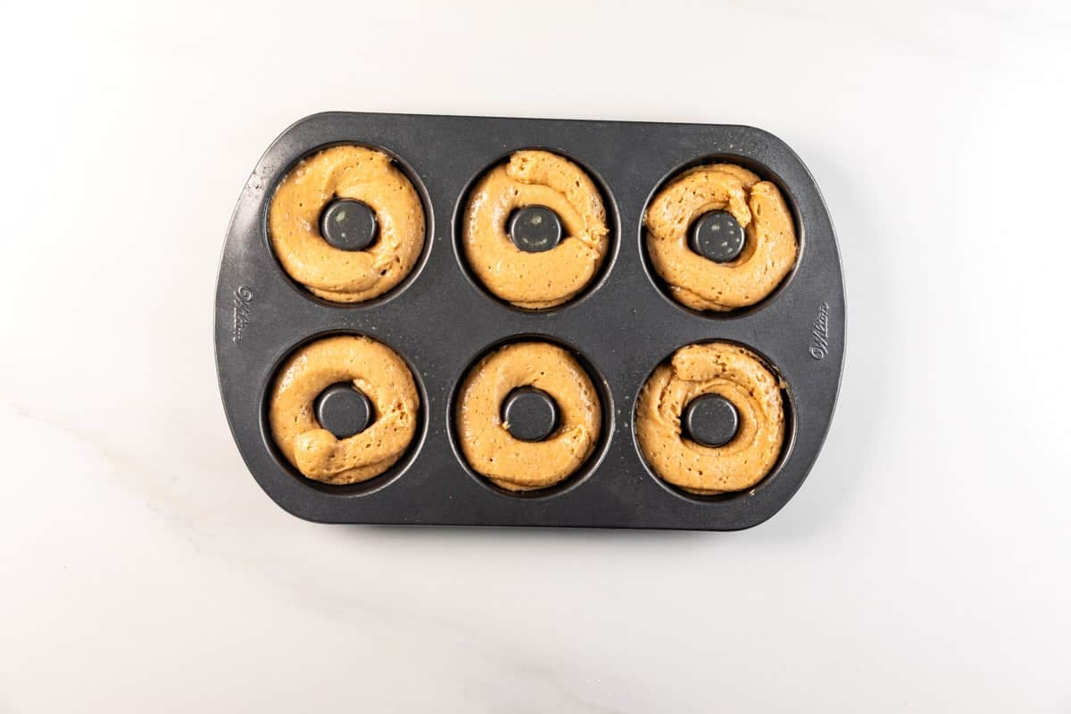 A black donut pan with six cavities, each filled with unbaked batter, sits on a white countertop.
