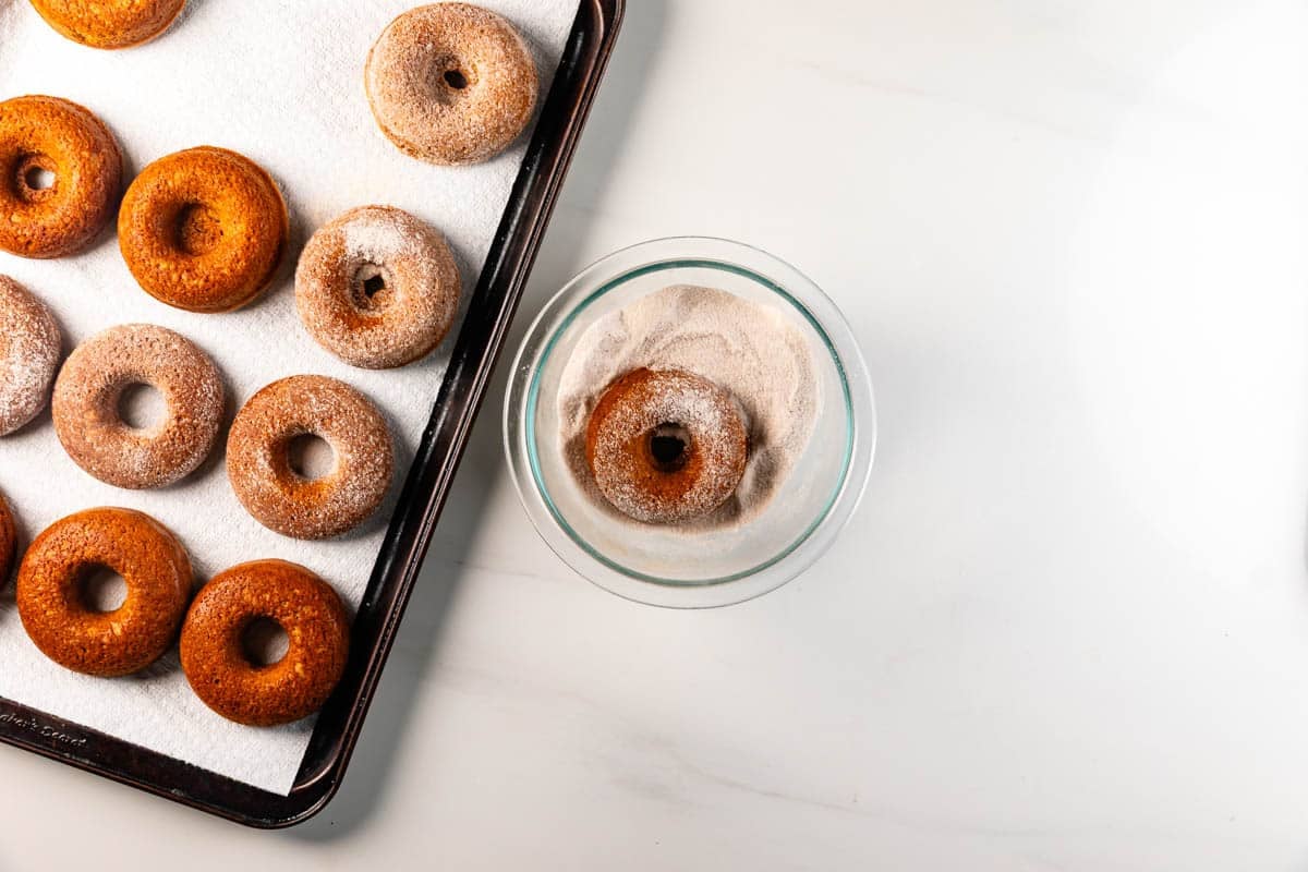 A tray of baked donuts rests on a paper towel, while one donut is being coated in cinnamon sugar in a glass bowl on a white surface.