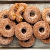 A metal tray filled with about a dozen sugar-coated baked donuts, some stacked and one partially broken, revealing the cake-like interior.