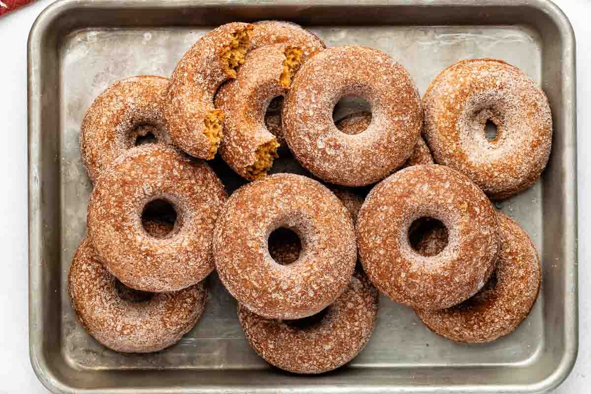 A metal tray filled with about a dozen sugar-coated baked donuts, some stacked and one partially broken, revealing the cake-like interior.