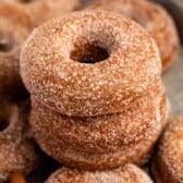 A stack of sugar-coated baked donuts sits on a tray, with more donuts and cinnamon sticks visible in the background.
