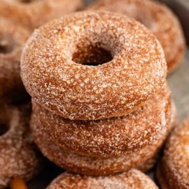 A stack of sugar-coated baked donuts sits on a tray, with more donuts and cinnamon sticks visible in the background.