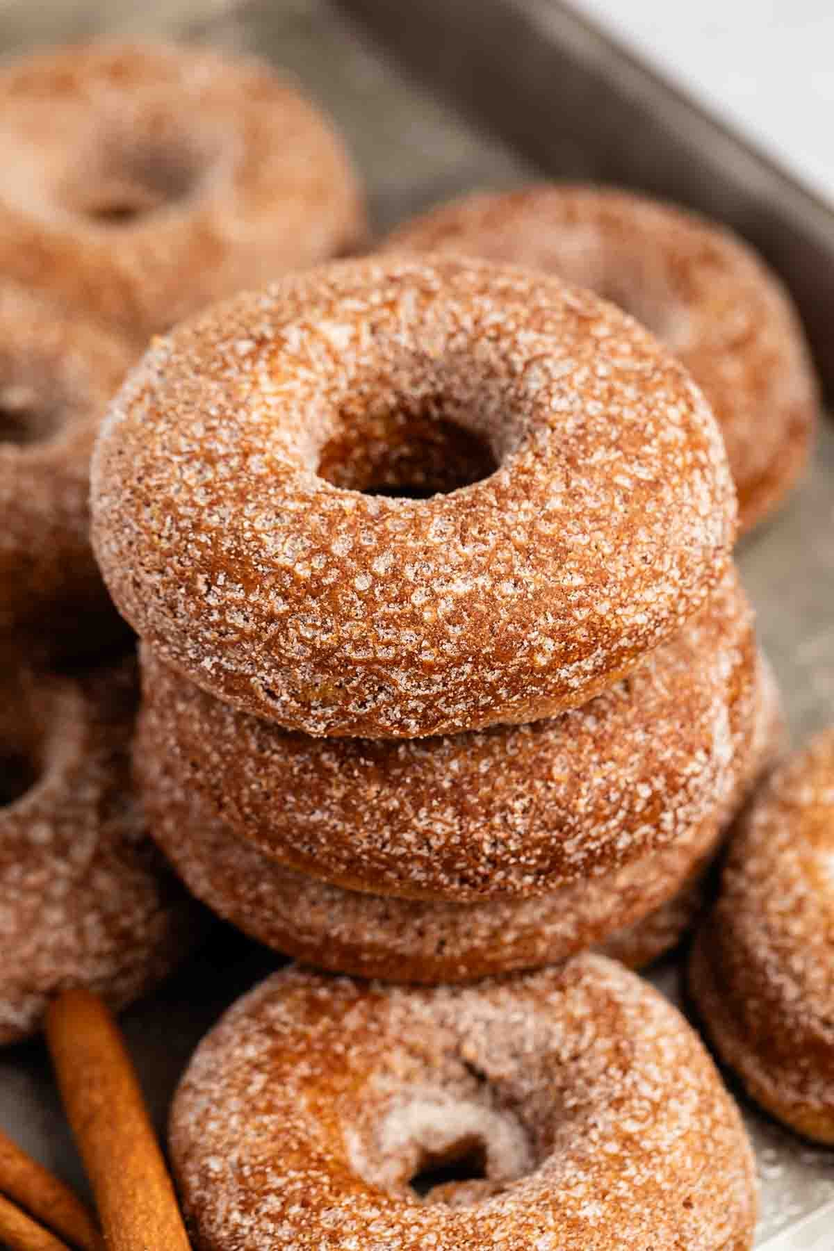 A stack of sugar-coated baked donuts sits on a tray, with more donuts and cinnamon sticks visible in the background.