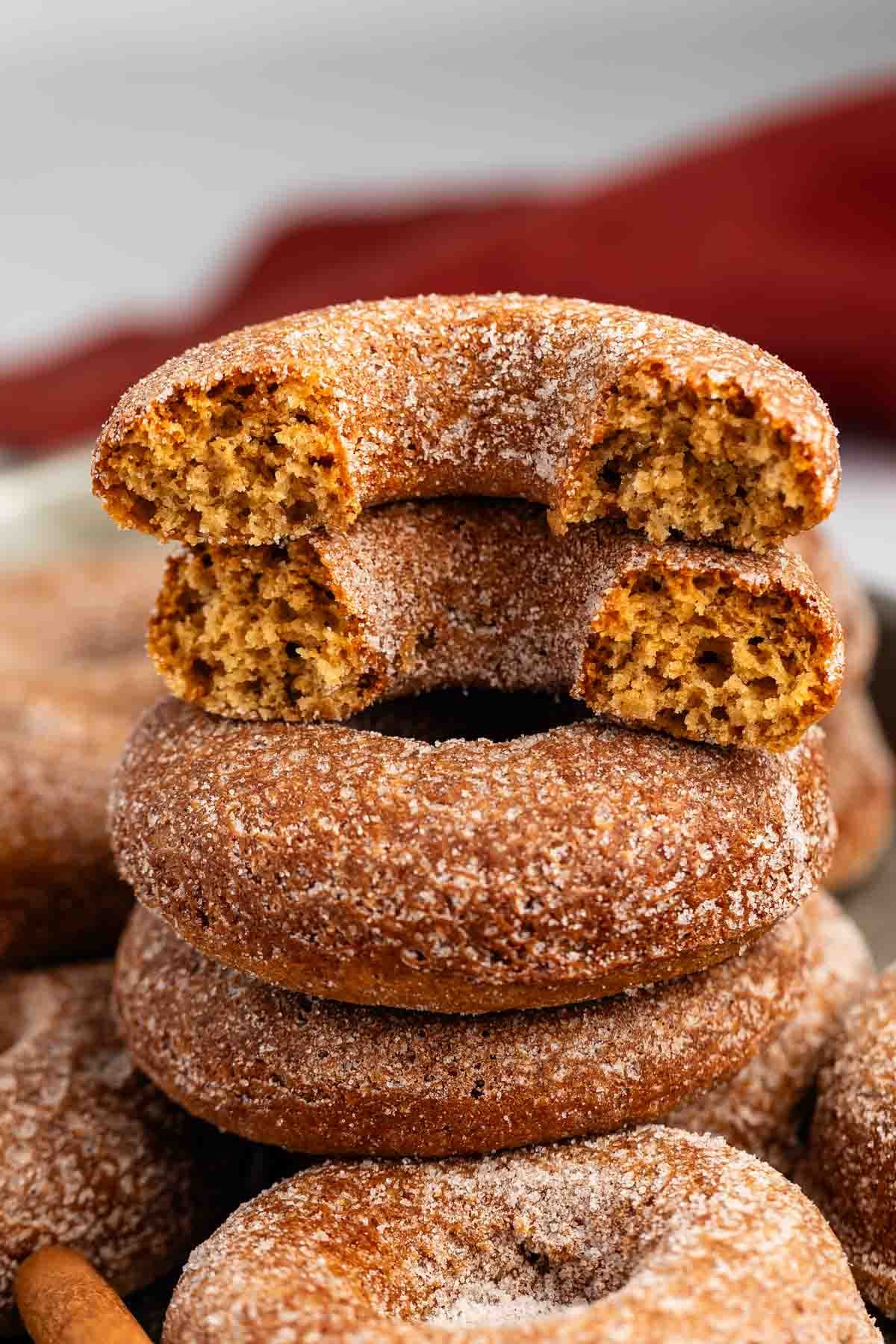 A stack of cinnamon sugar donuts, with one donut on top broken in half to show its soft, fluffy inside. The donuts are coated in a layer of sugar and arranged against a blurred background.