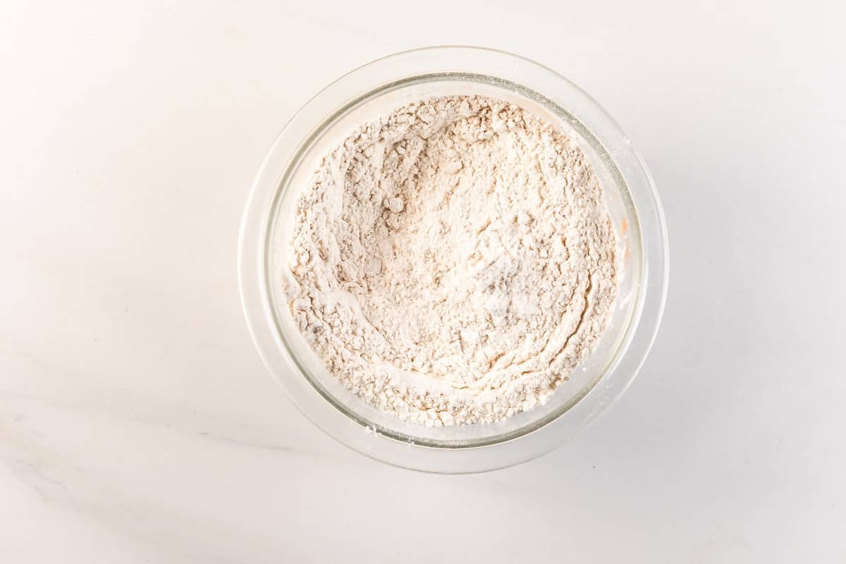 A clear glass bowl filled with white flour sits on a light-colored marble surface, viewed from above.