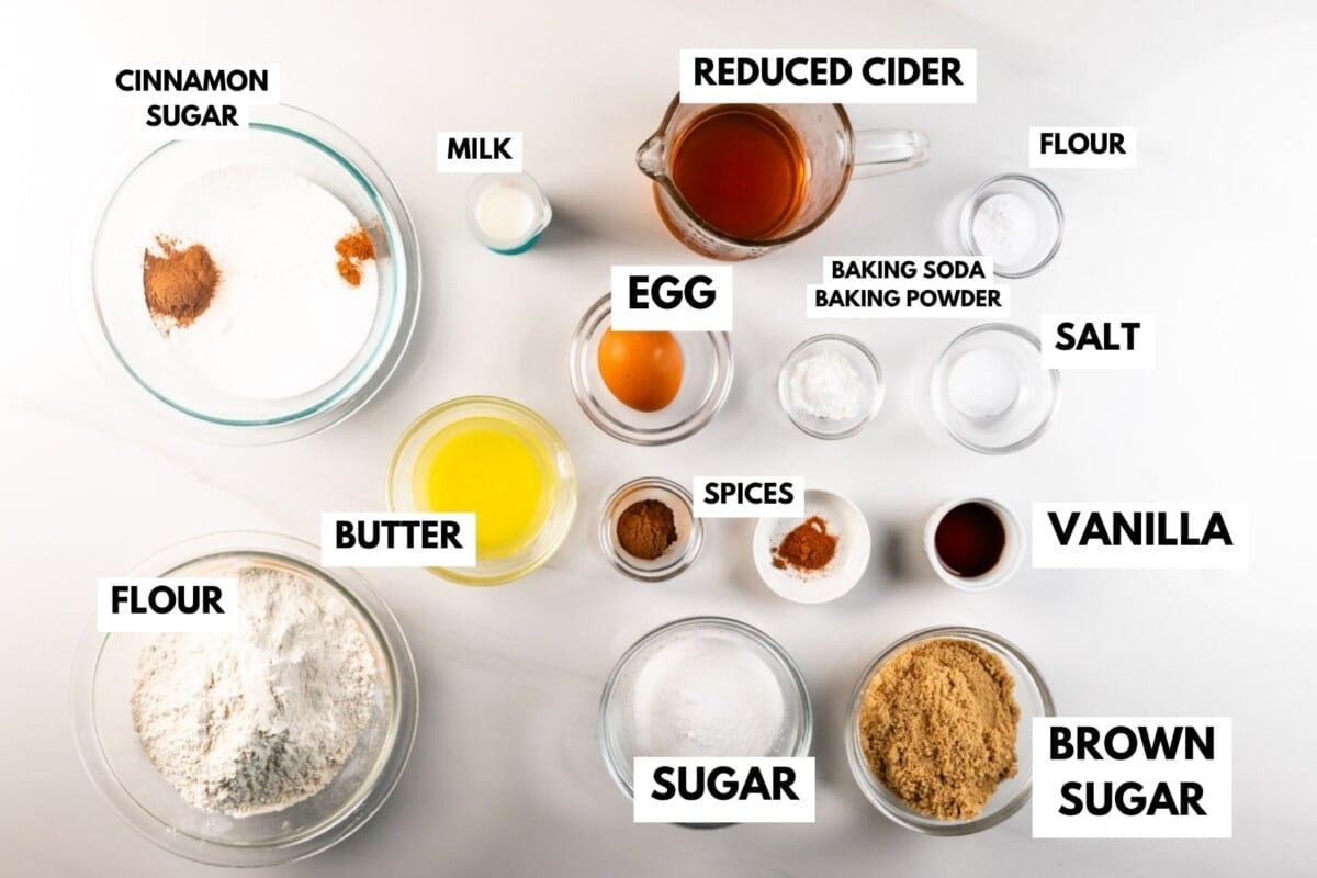 Baking ingredients on a white surface, each labeled: cinnamon sugar, milk, reduced cider, flour, egg, butter, sugar, brown sugar, baking soda, baking powder, salt, vanilla, and spices.