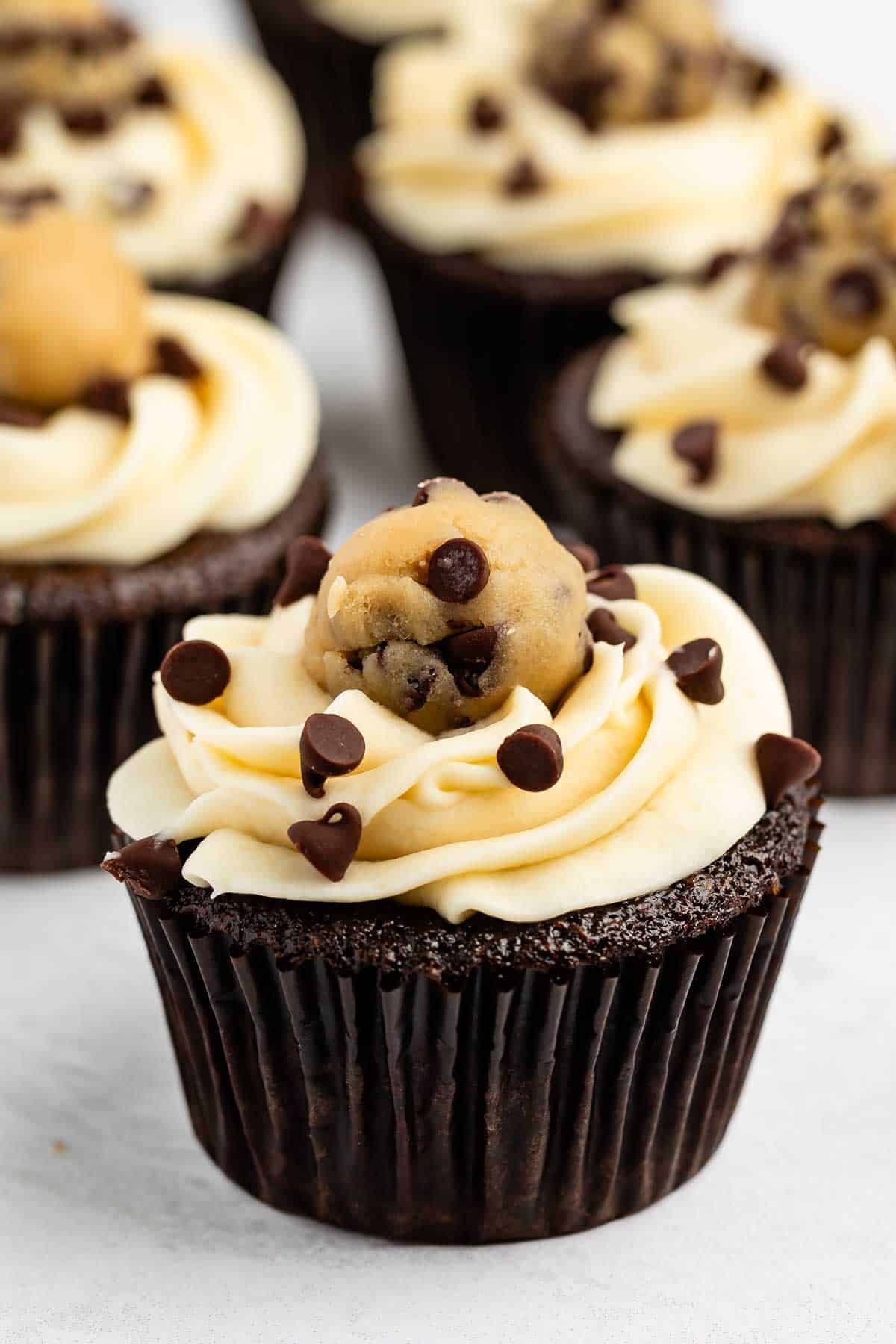Chocolate cupcake with vanilla frosting, topped with chocolate chips and a ball of cookie dough. Several similar cupcakes are blurred in the background on a white surface.