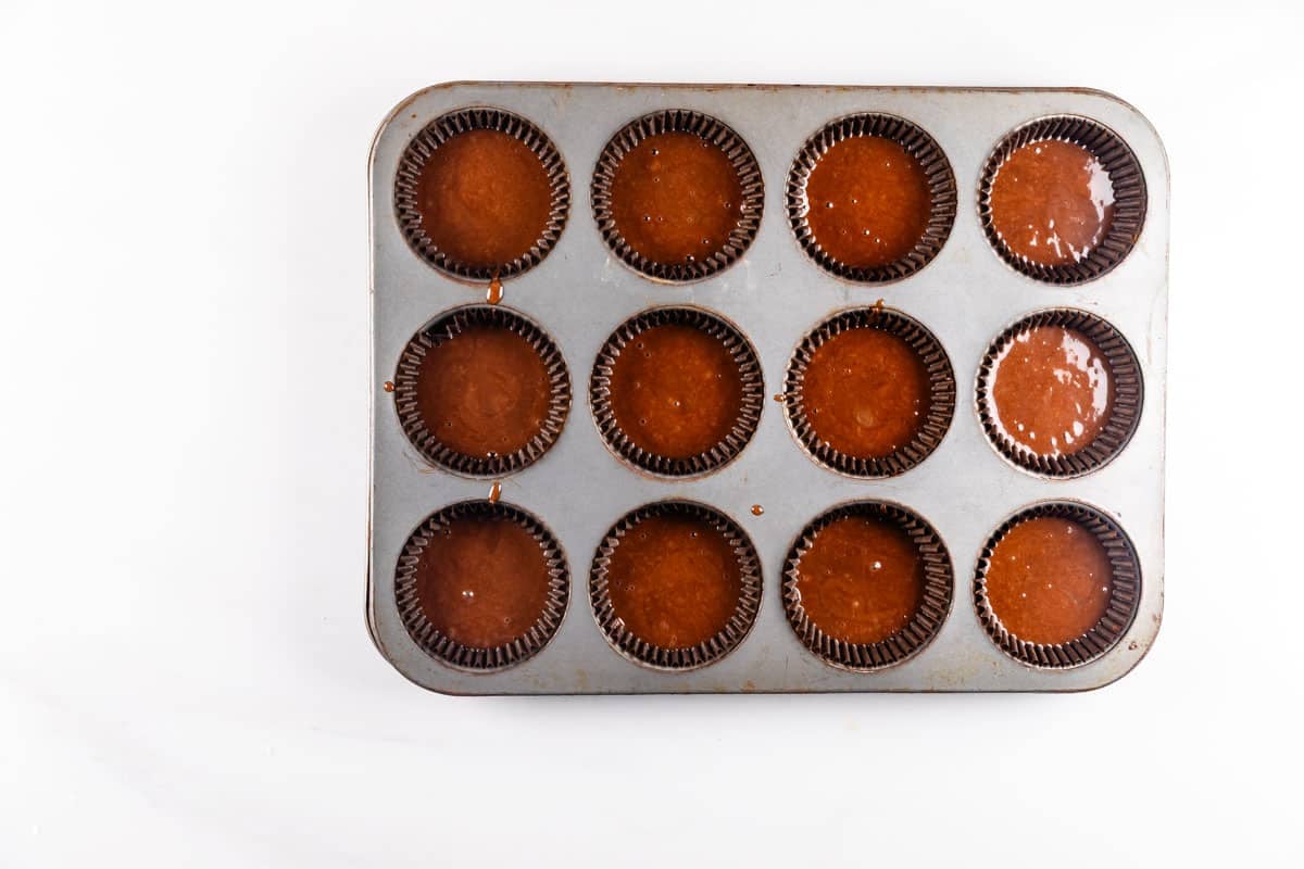 A muffin tin with twelve cups, each lined with brown paper liners, filled with chocolate batter, ready to be baked. The tin sits on a white surface.