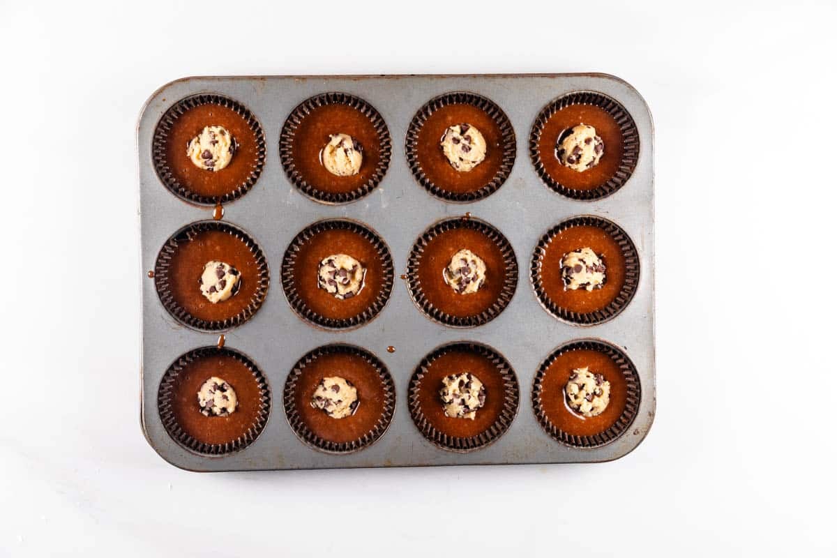 A metal muffin tray with twelve chocolate cupcakes, each topped with a small dollop of chocolate chip cookie dough, seen from above on a white background.