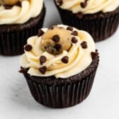 Chocolate cupcake with vanilla frosting, topped with a ball of chocolate chip cookie dough and sprinkled with mini chocolate chips, on a white surface.
