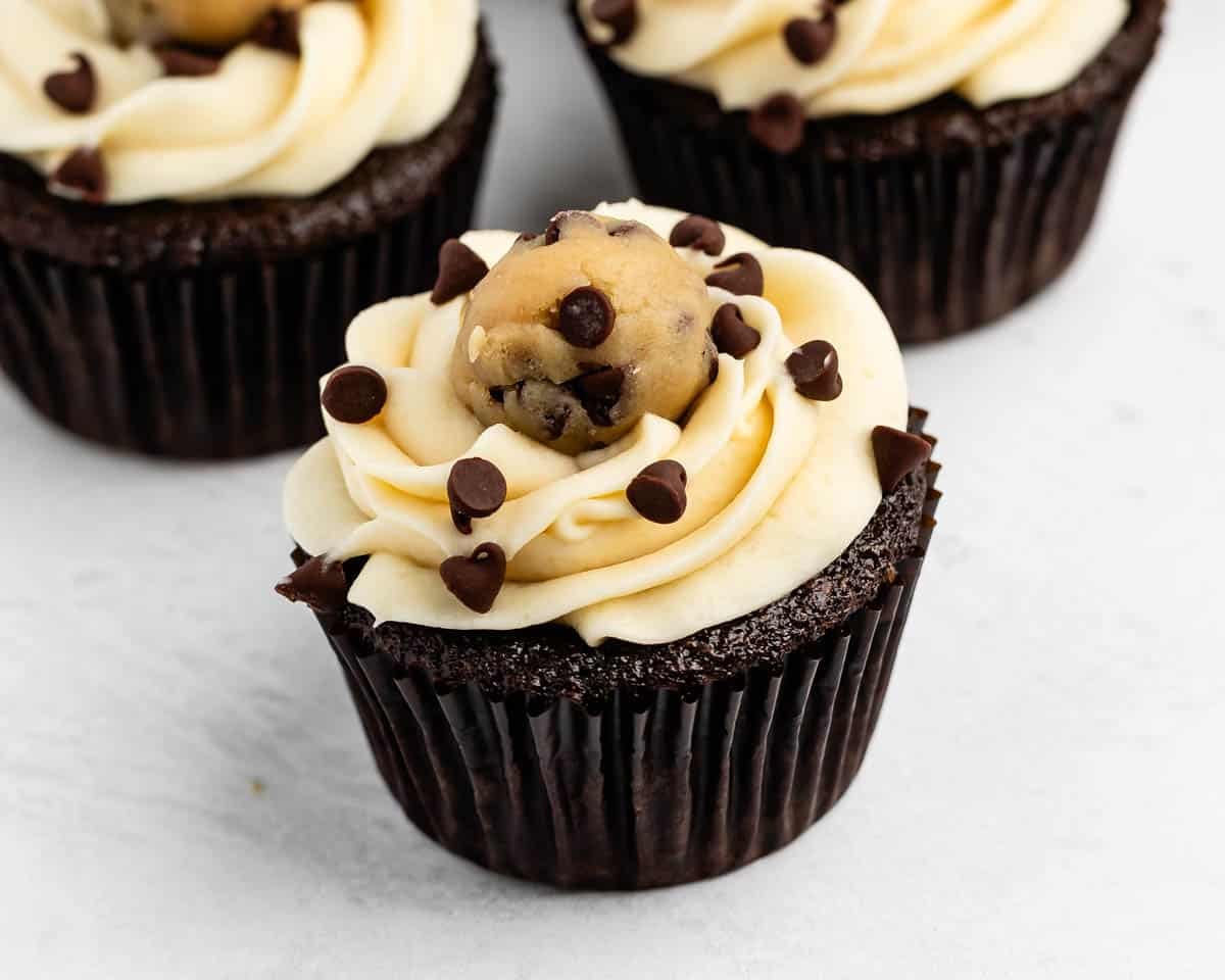 Chocolate cupcake with vanilla frosting, topped with a ball of chocolate chip cookie dough and sprinkled with mini chocolate chips, on a white surface.